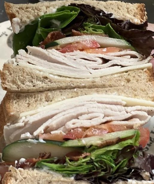 Tall stacked turkey sandwich with bacon, cheese, tomato, cucumber, greens on wheat bread